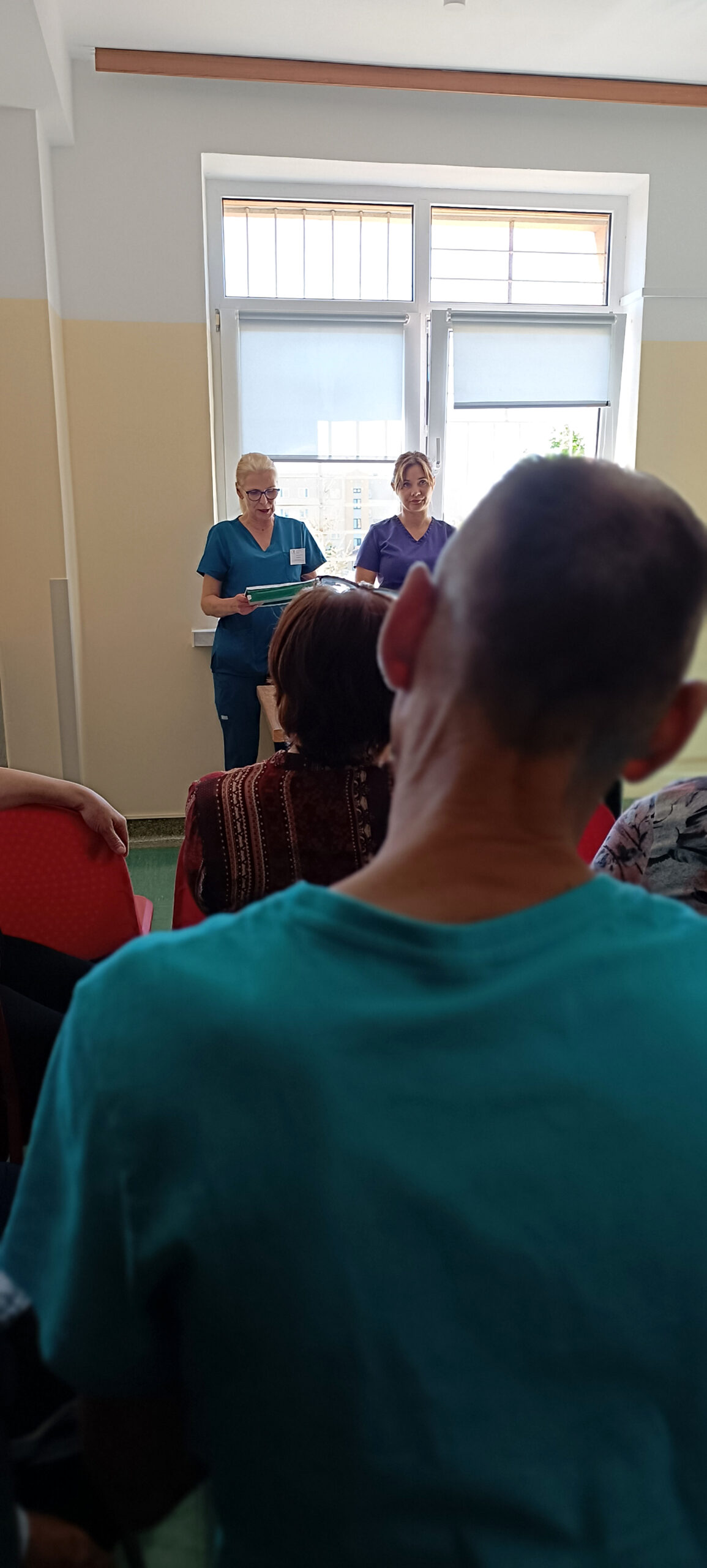 Wykład na oddziale geriatrycznym Dwie panie stoją pod oknem i prowadzą wyklad Grupa ludzi słucha
