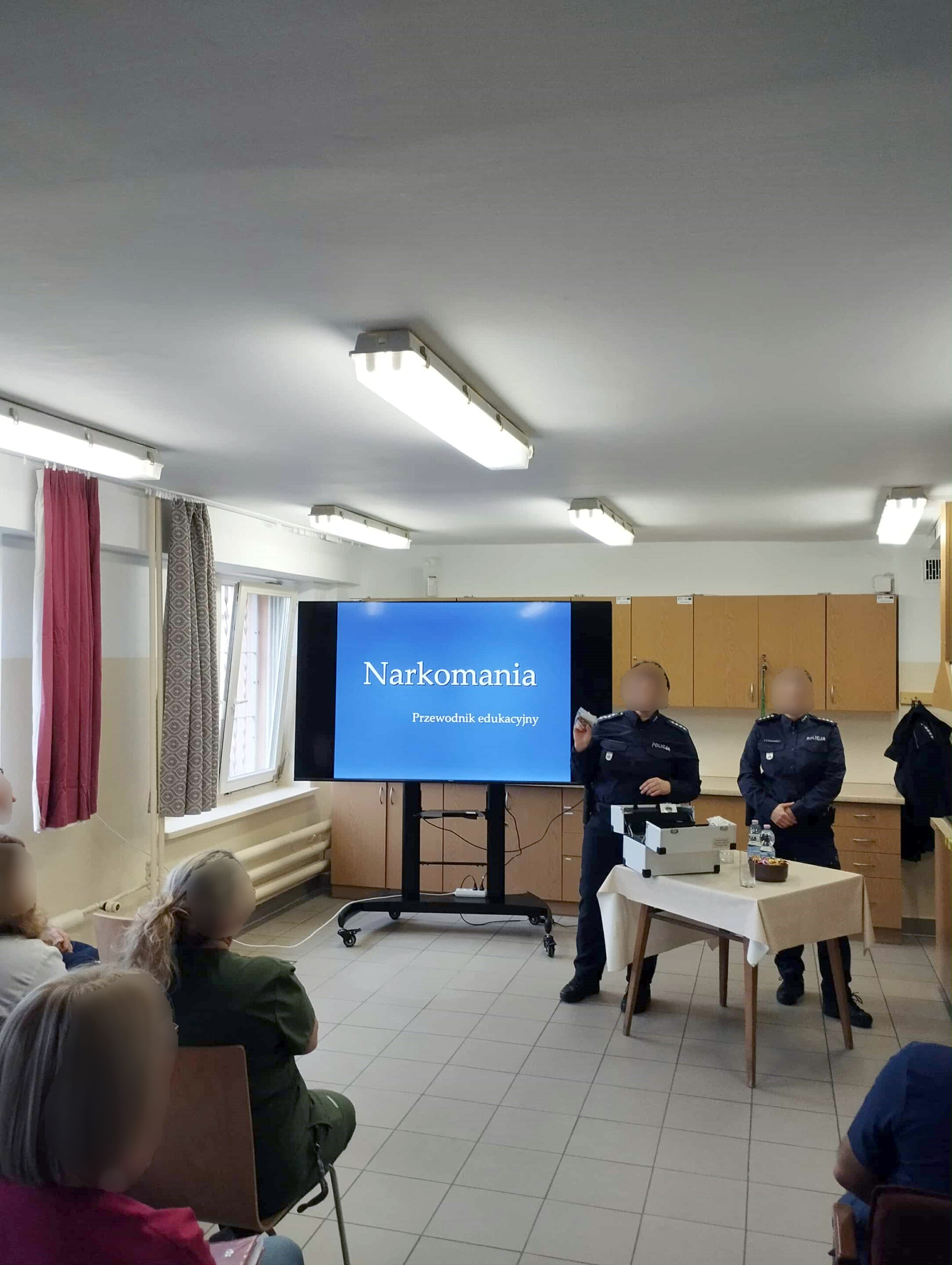 Dwie Panie policjantki w mundurach bez czapek szkolą personel. Oświetlona sala w dzień z tyłu duży telewizor z opisem szkolenia