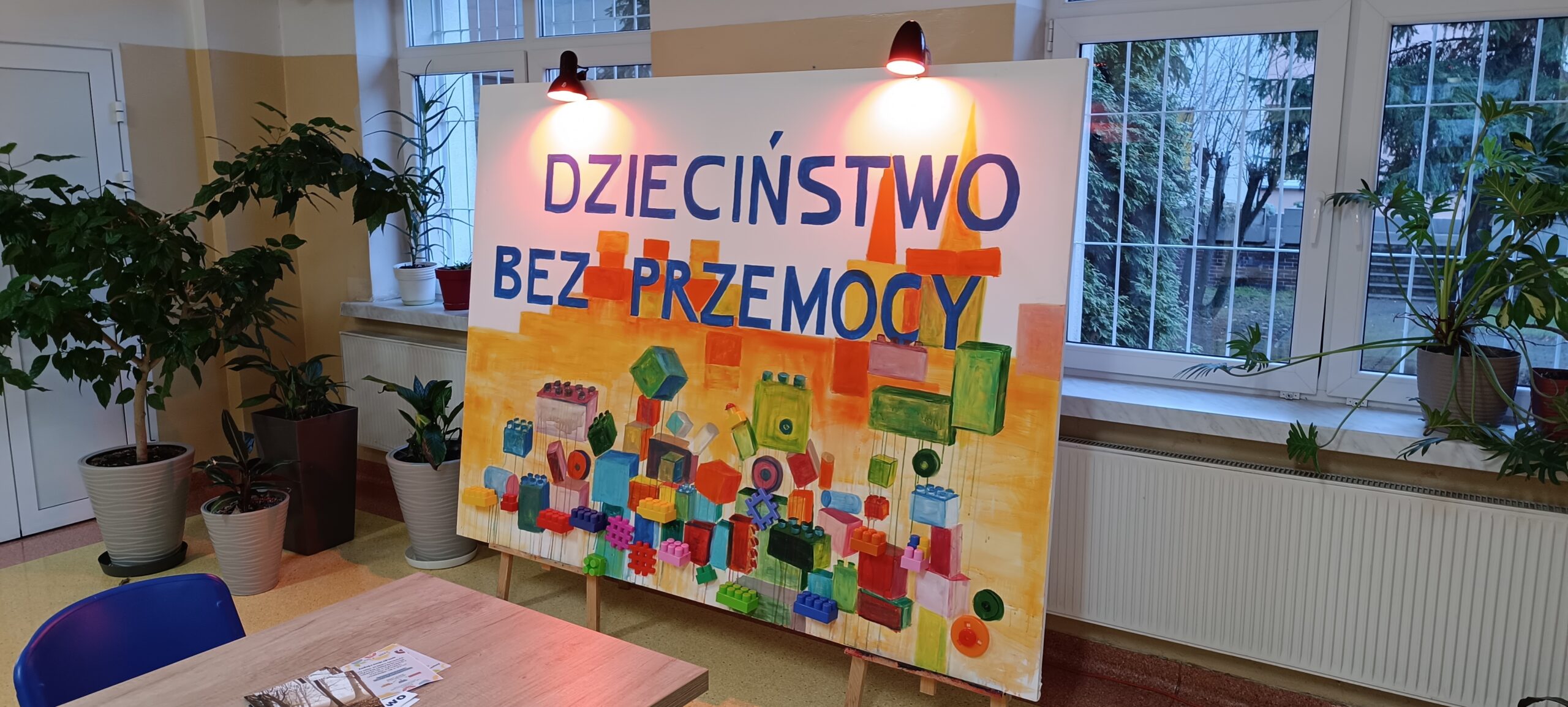 Duży plakat stojący na korytarzu , podświetlony na czerwono o tematyce przemocy wobec dzieci. Plakat przedstawia rozsypane klocki i duży napis Dzieciństwo Bez Przemocy