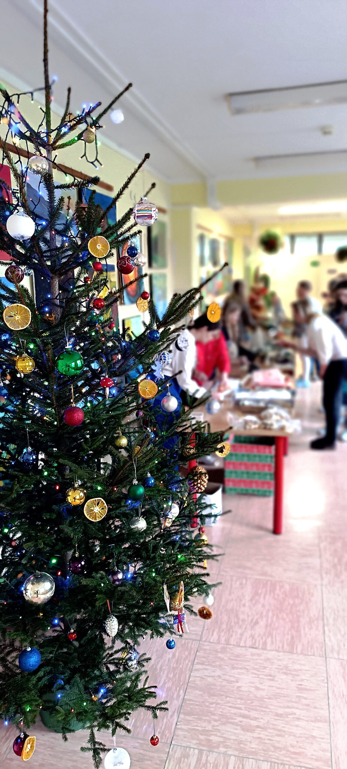 Grupa ludzi ogląda stroiki i ozdoby świateczne wystawione na szkolnych ławkach w korytarzy szpitala