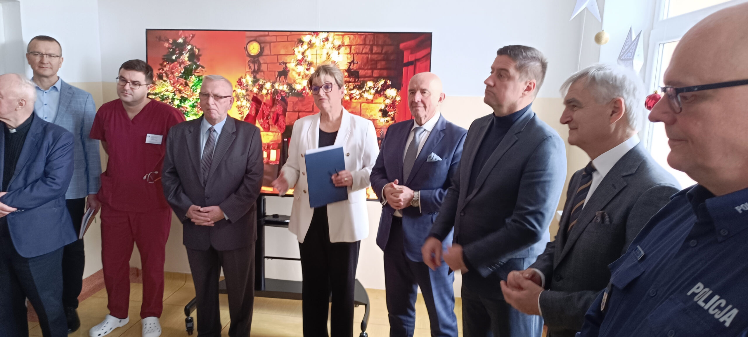 Grupa Ludzi na spotkaniu opłatkowym w dużej sali konferencyjnej składa sobie życzenia, 3 stoły pełne potraw wigilijnych i czerwonego barszczu