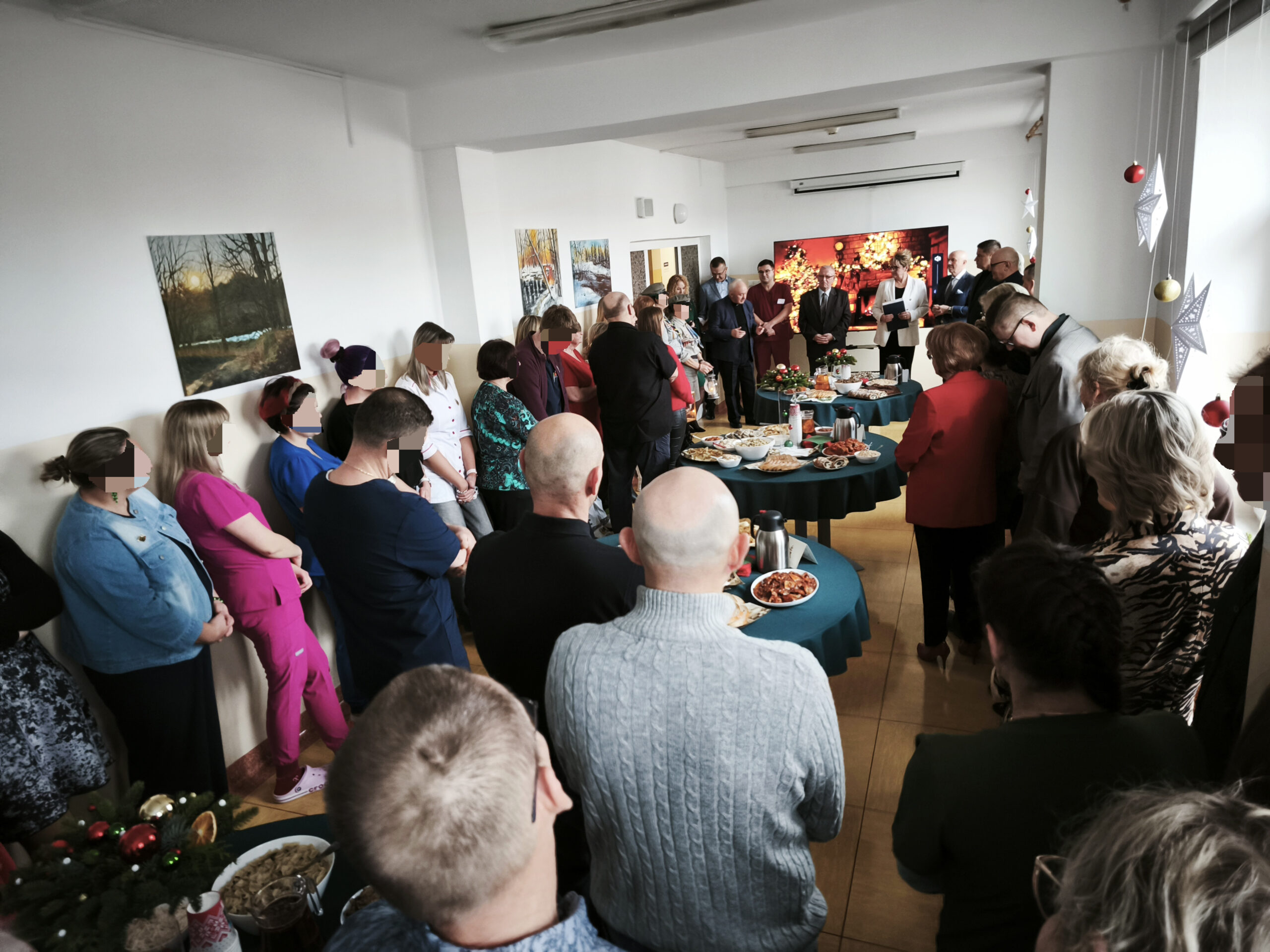 Grupa Ludzi na spotkaniu opłatkowym w dużej sali konferencyjnej składa sobie życzenia, 3 stoły pełne potraw wigilijnych i czerwonego barszczu