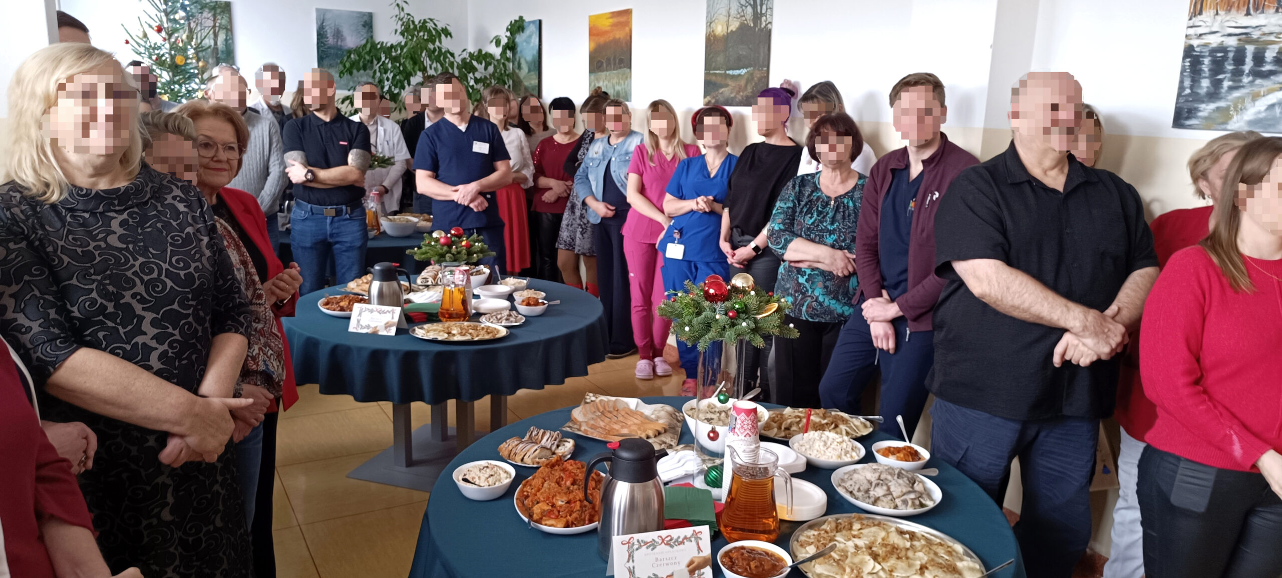 Grupa Ludzi na spotkaniu opłatkowym w dużej sali konferencyjnej składa sobie życzenia, 3 stoły pełne potraw wigilijnych i czerwonego barszczu