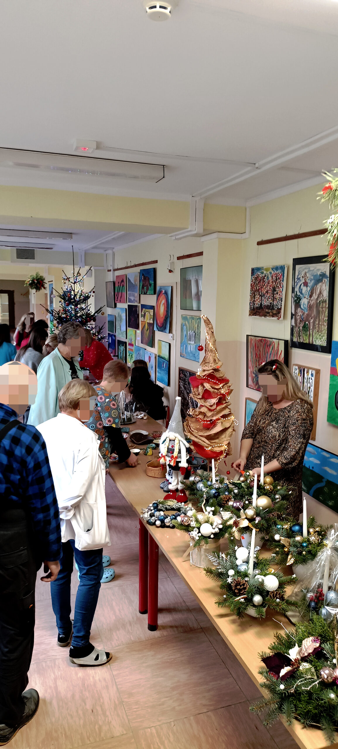 Grupa ludzi  ogląda stroiki i ozdoby świateczne wystawione na szkolnych ławkach w korytarzy szpitala