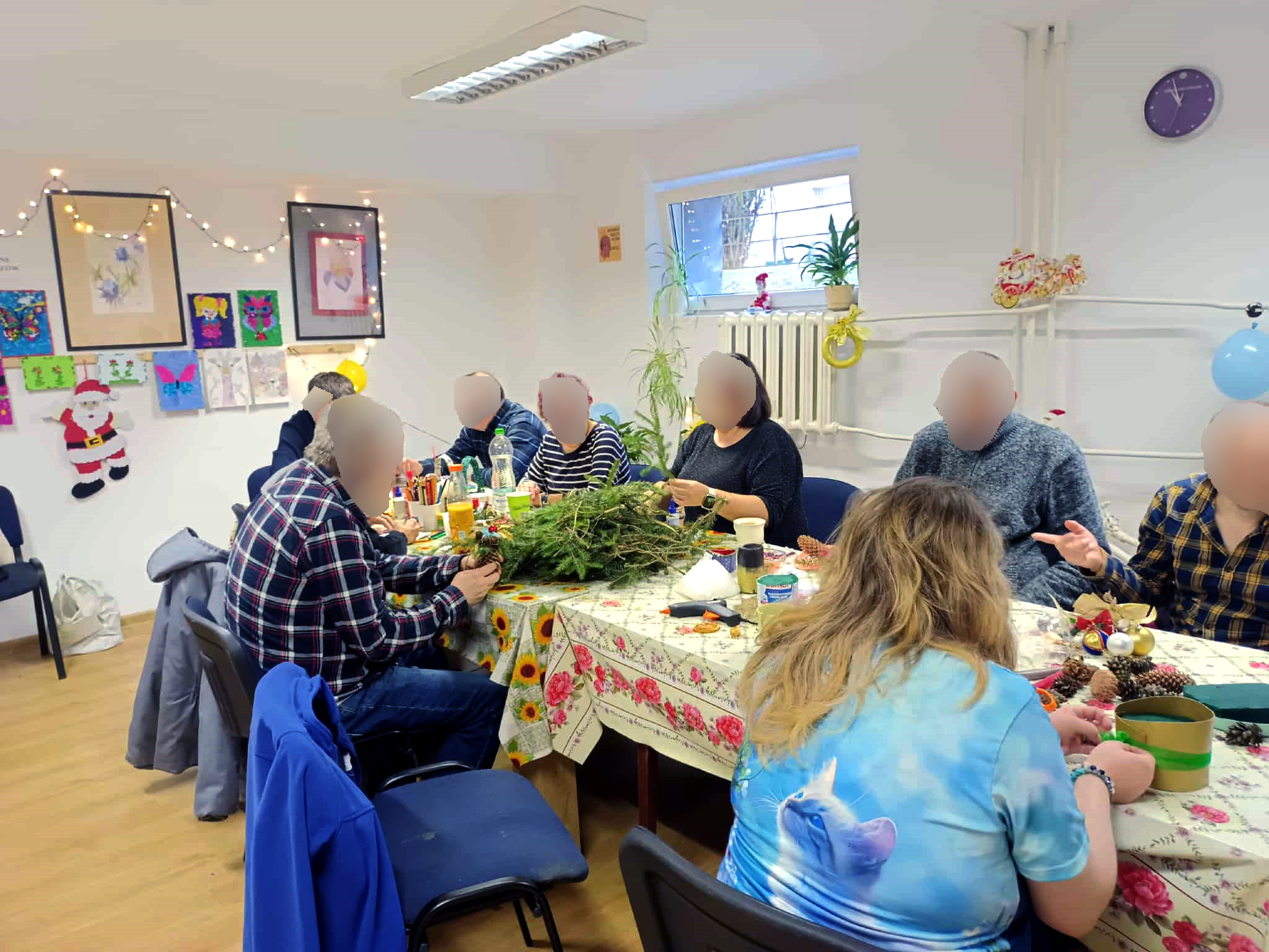 Zdjęcie przedstawia salę, na której stoi stół a na nim stroiki świąteczne przygotowane przez osoby z klubu pacjenta. Wokół stołu siedzą osoby, mężczyźni i kobiety z klubu pacjenta