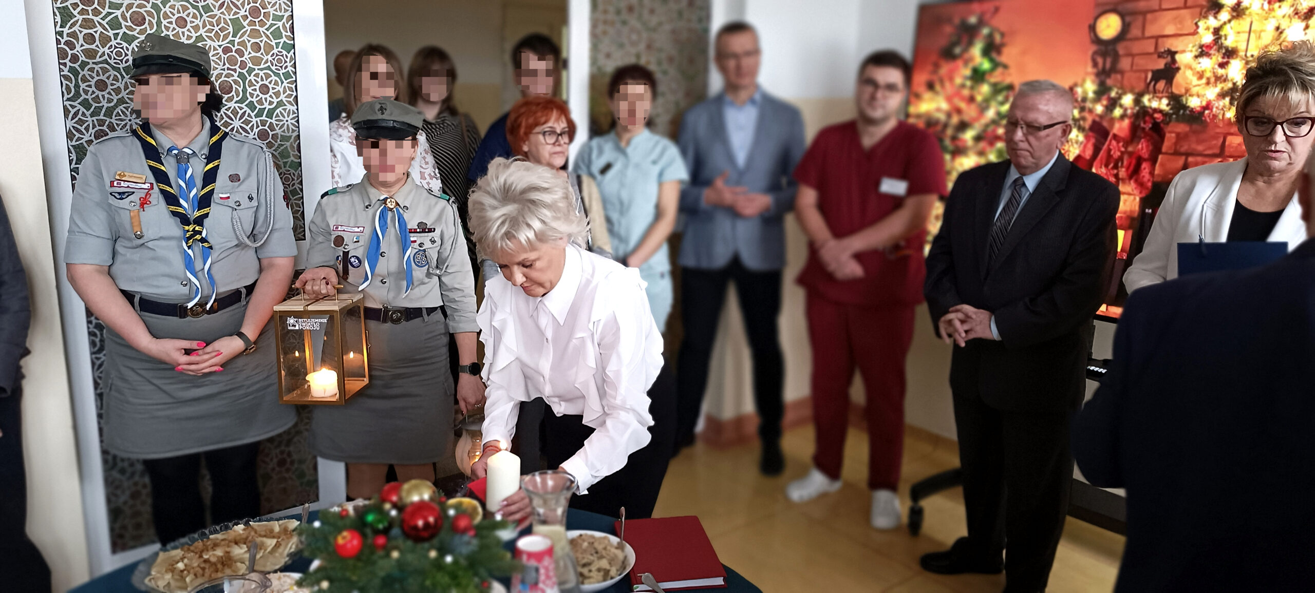 Grupa Ludzi na spotkaniu opłatkowym w dużej sali konferencyjnej składa sobie życzenia, 3 stoły pełne potraw wigilijnych i czerwonego barszczu