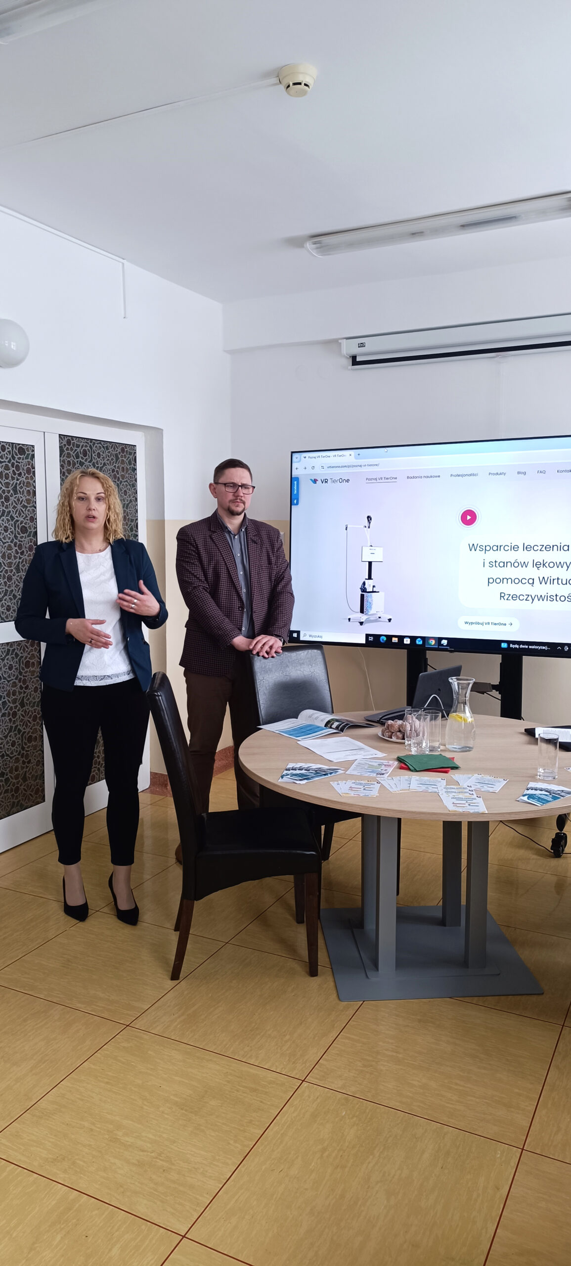 Mężczyzna i kobieta na sali konferencyjnej prowadzą wykład, w tle duży telewizor z wyświetlonymi materiałami