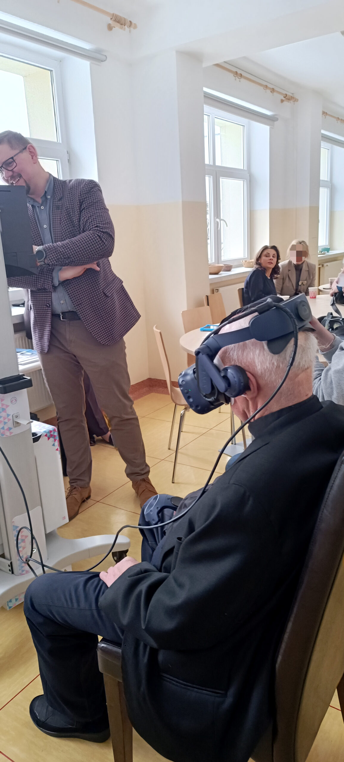 mężczyzna w sutannie z założonymi goglami wirtualnymi na głowie siedzi w sali konferencyjnej