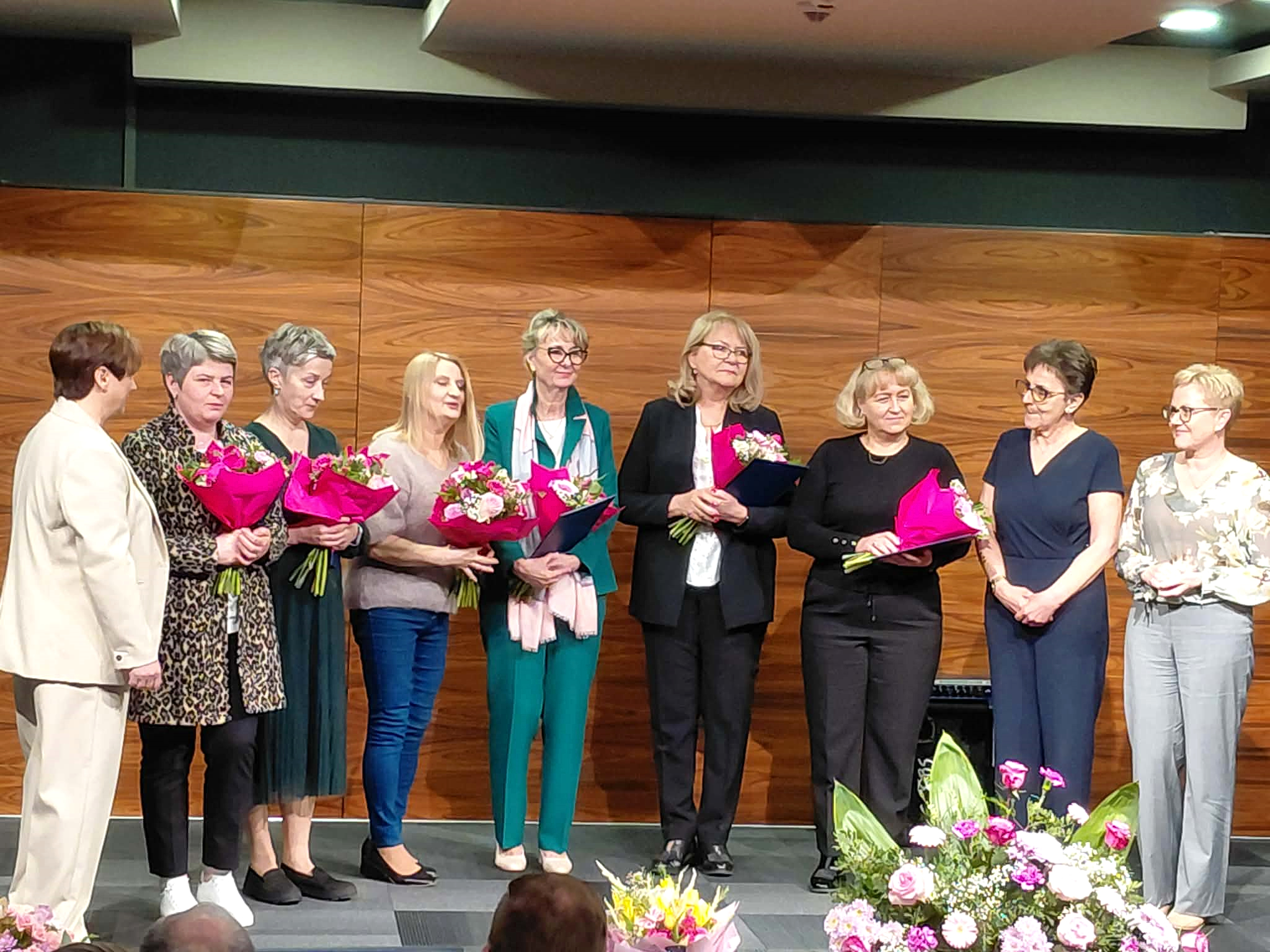 Sala konferencyjna , wręczenie nagród. Grupa kobiet stoi w półkolu na scenie z kwiatami i wręczonymi wyróżnieniami