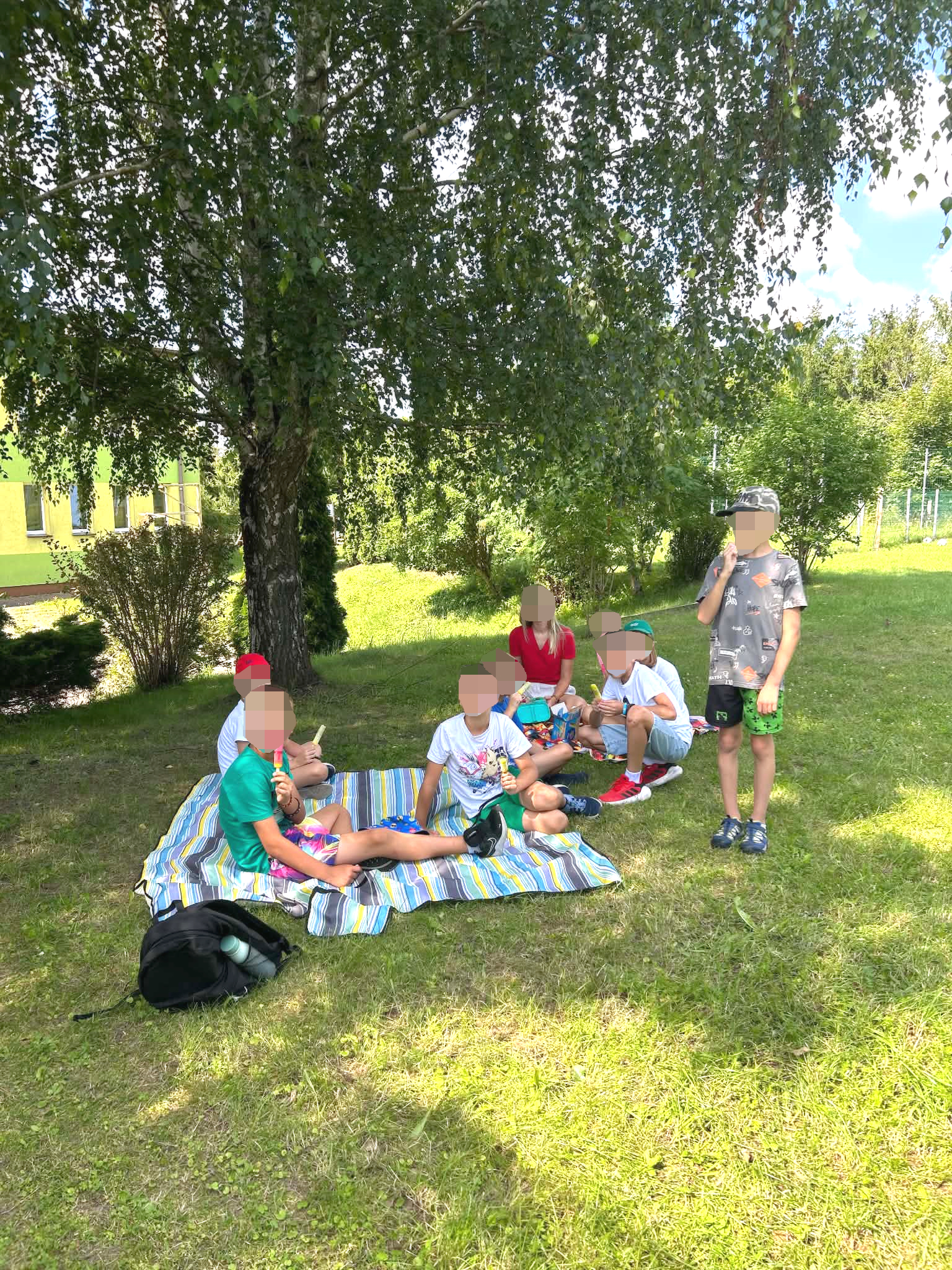 Grupa dzieci siedzi na kocu w pięknej słonecznej atmosferze na polanie w tle drzewa i stara szkoła